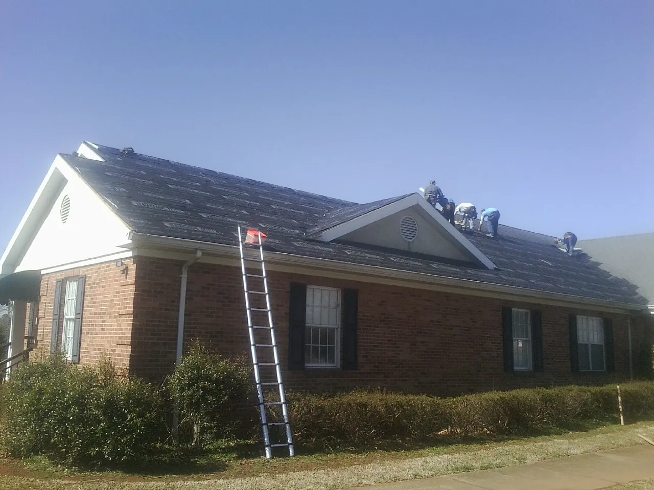 Crew tearing shingles off a brick ranch roof with a ladder set — Metal Roof Repair work by our crew in Glen Cove