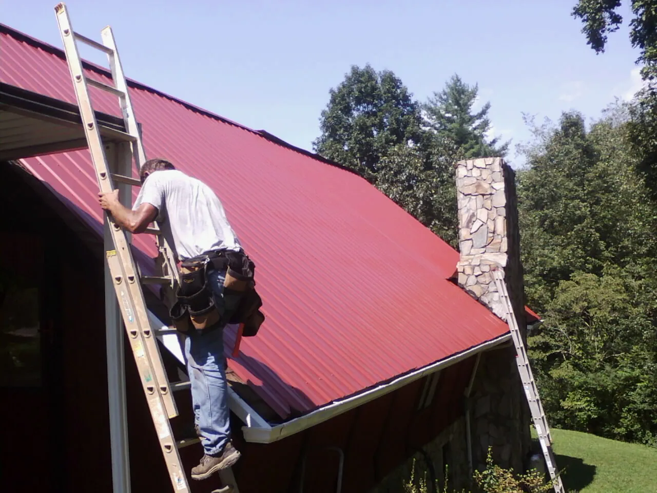 Roofer on a ladder beside a red metal roof and stone chimney — Soffit & Fascia Repair by Roofing Brothers in Glen Cove
